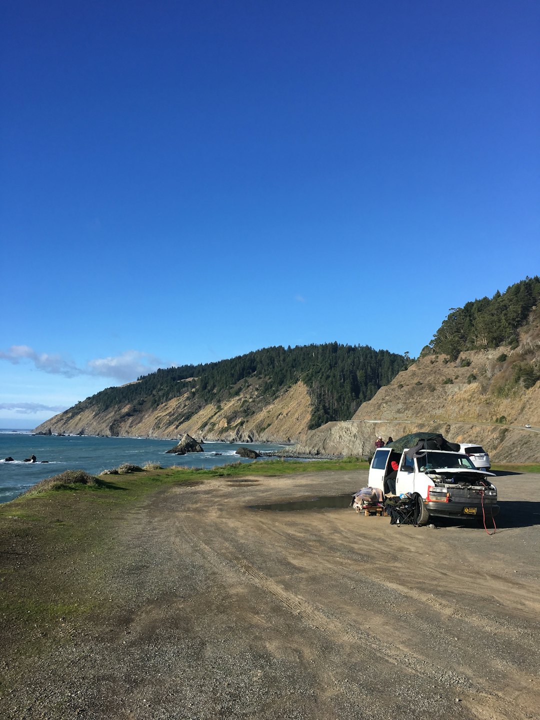 A white van with an open hood parked near a coastal road.